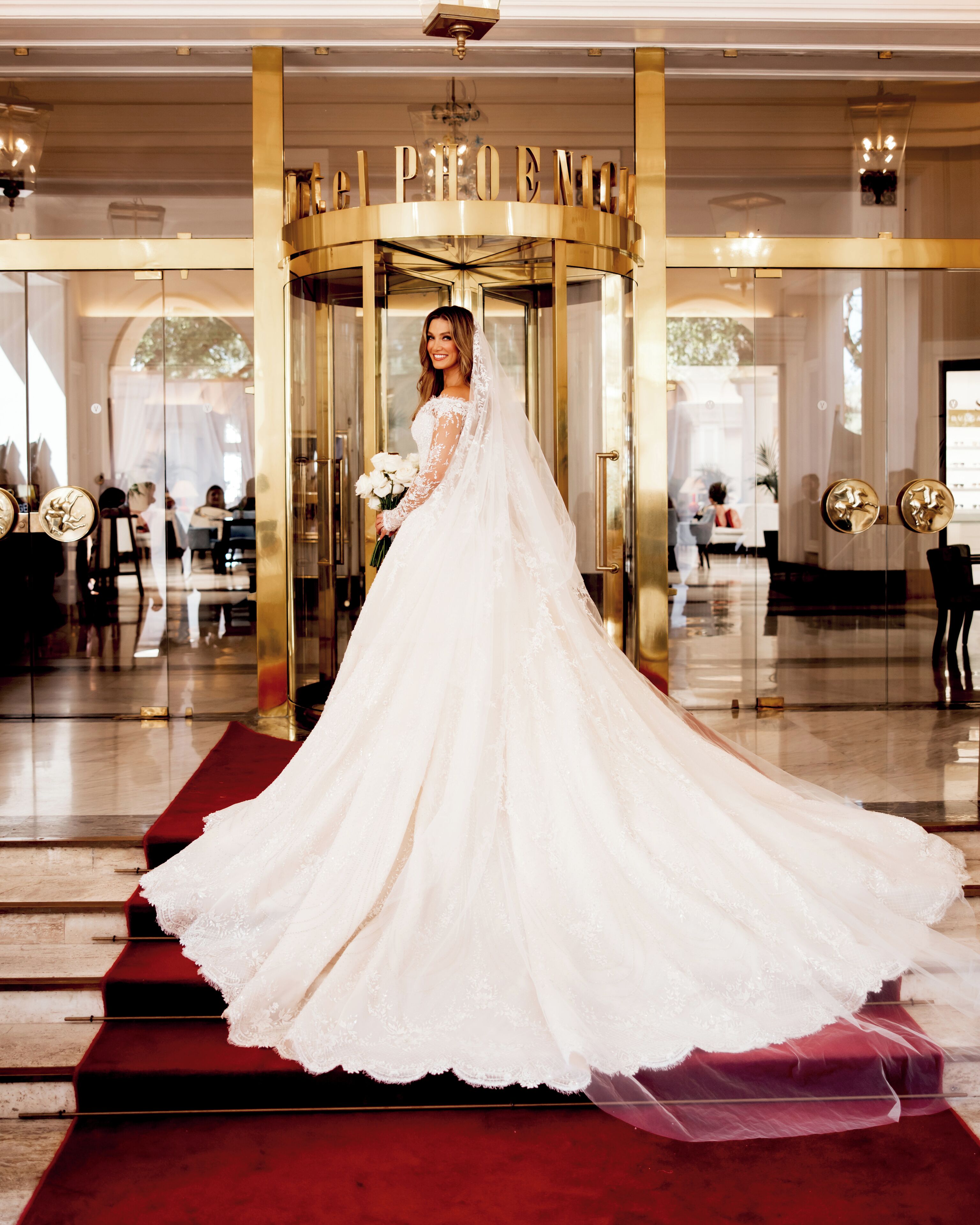 A bride in a white wedding dress standing at the entrance of a hotel with a red carpet.
