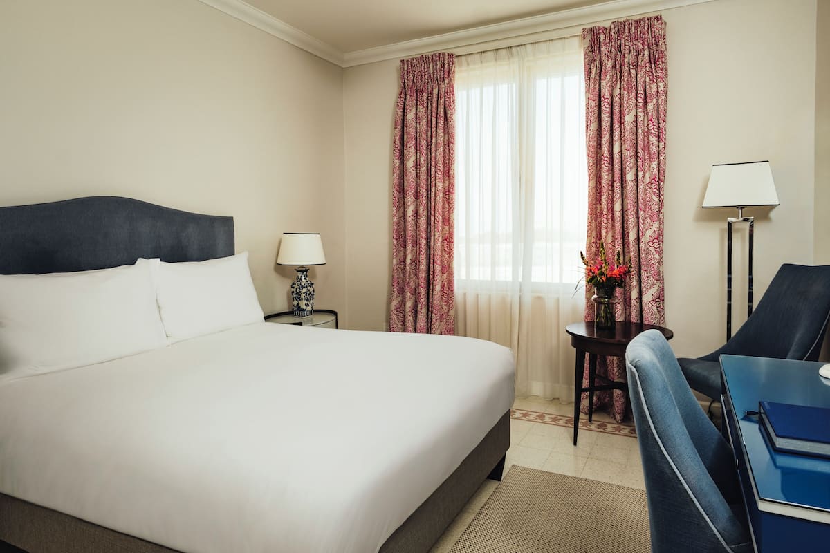 A hotel room with a bed, a desk, a chair, a lamp, and a window with curtains.