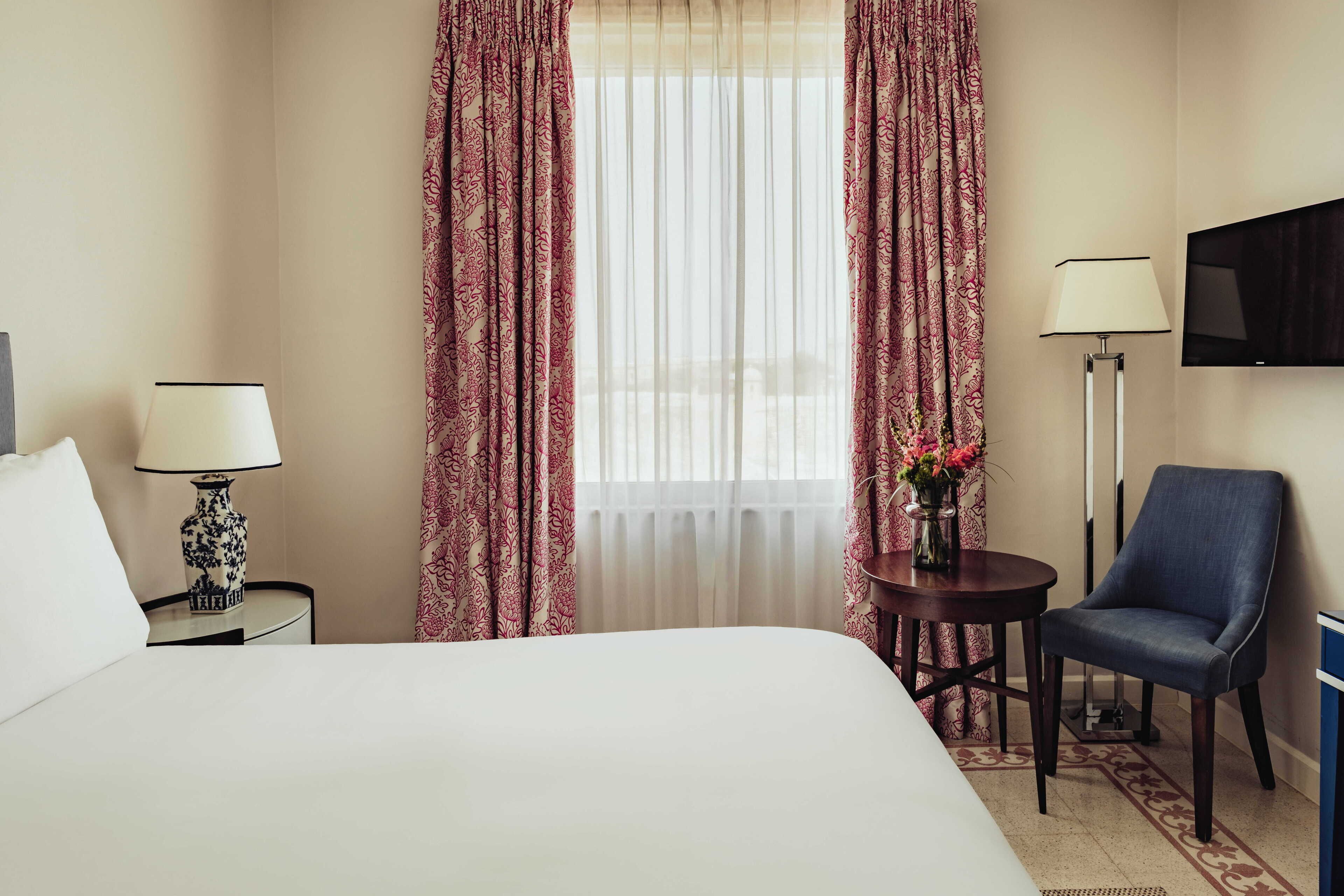 Habitación de hotel con una cama, dos mesitas de noche con lámparas, una silla, una mesita con un jarrón, un televisor montado en la pared y una ventana con cortinas traslúcidas.
