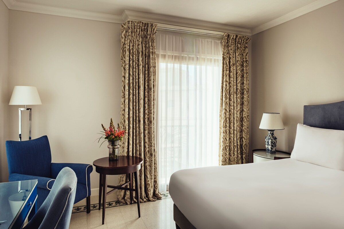 Habitación de hotel con cama, escritorio con lámpara, silla y ventana con cortinas.