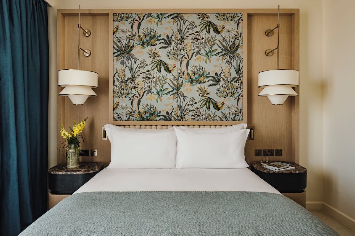 A hotel room with a bed, two bedside lamps, a nightstand with a vase of flowers, and a wall with a floral pattern.