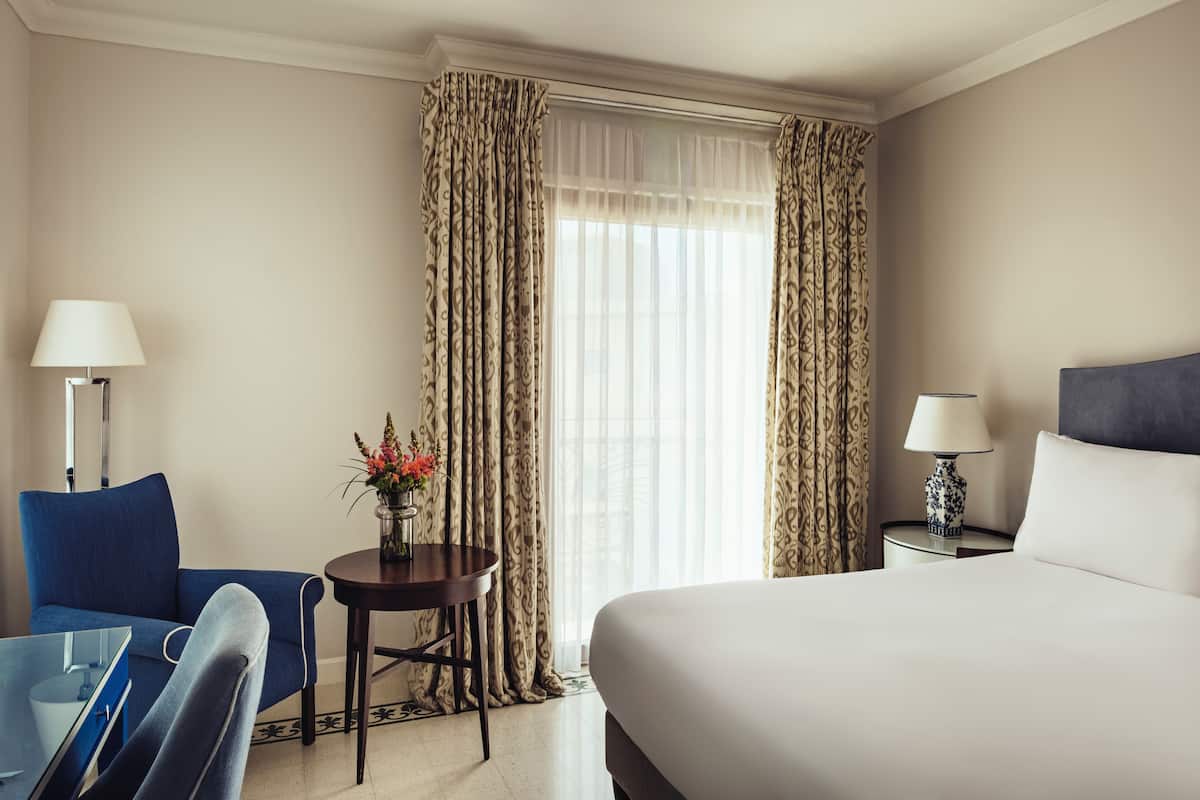 A hotel room with a bed, a blue armchair, a small table with a vase of flowers, and curtains.