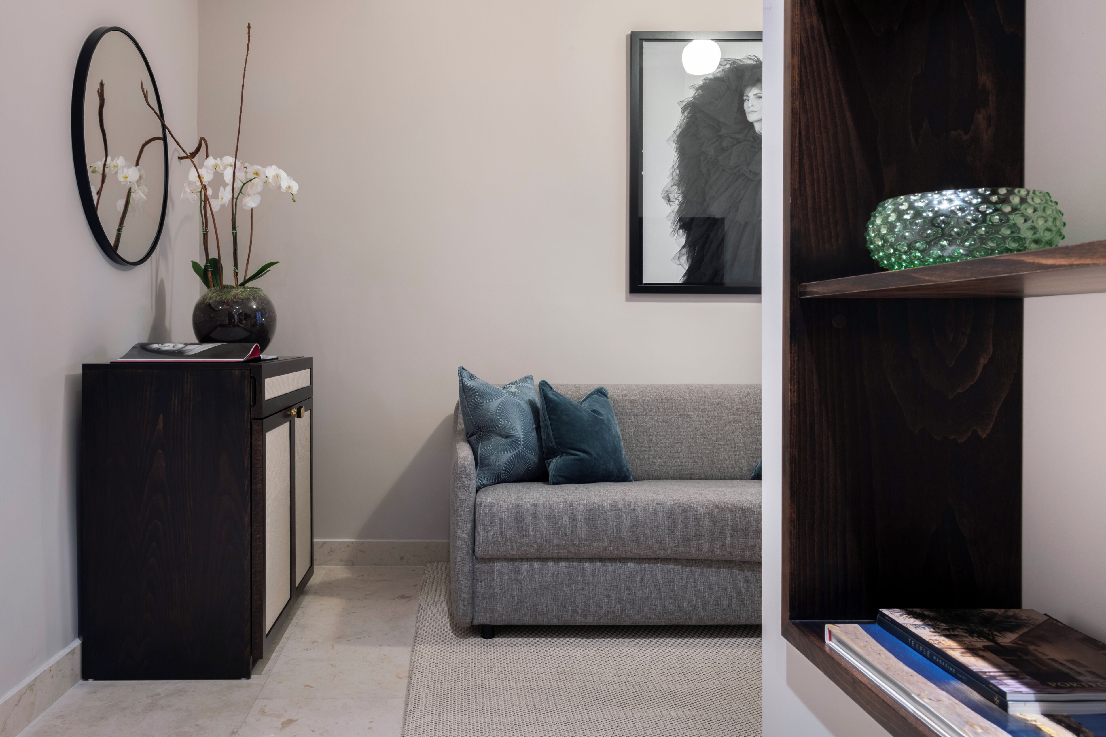 A room with a grey sofa, a dark wooden cabinet, a vase with white flowers, a framed artwork, and a green decorative item on a shelf.