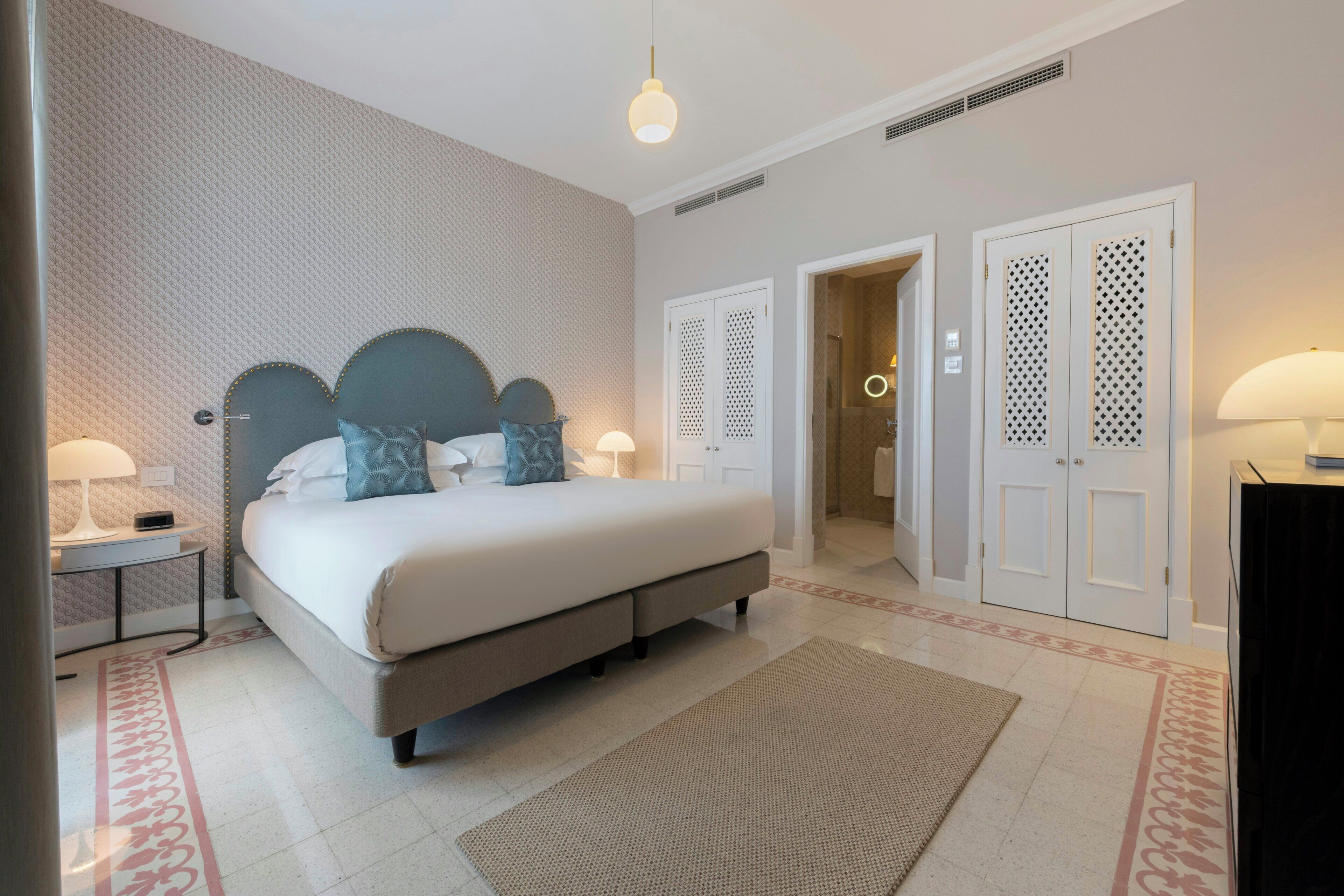A bedroom with a bed, nightstands, a lamp, a rug, and a door leading to another room.