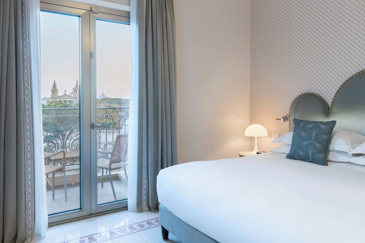 A hotel room with a bed, a nightstand, a lamp, a view of a balcony with chairs, and a patterned wall.