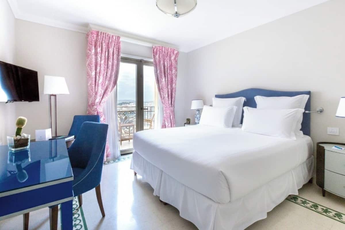 A hotel room with a bed, a desk with a chair, a TV, and a balcony with pink curtains.