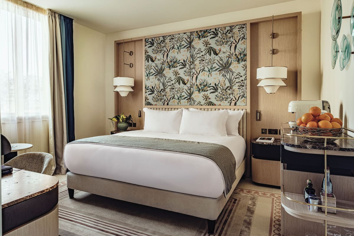 A hotel room with a large bed, a desk, a chair, a nightstand, and a wall-mounted lamp.