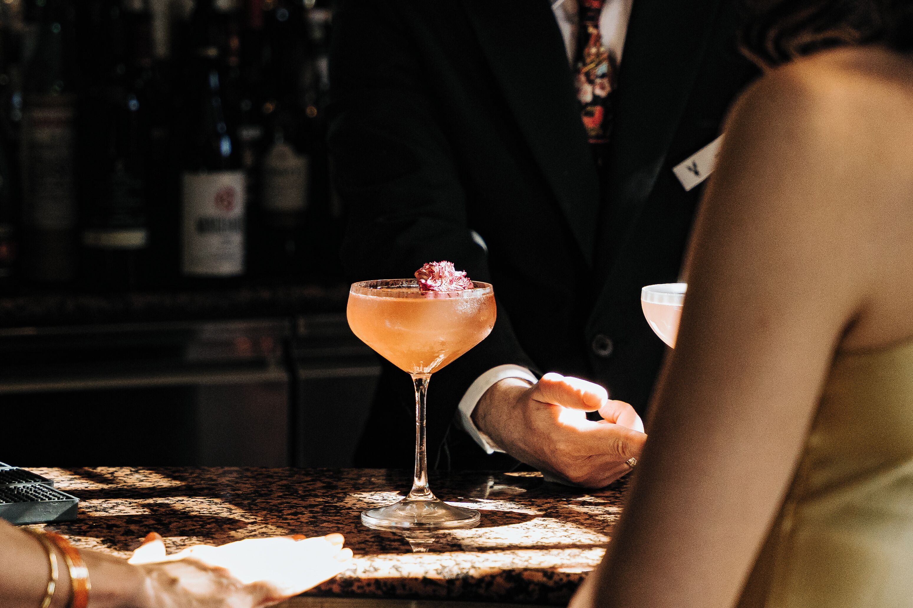 A bar setting with a cocktail in a glass, a person in a suit, and a bar counter.
