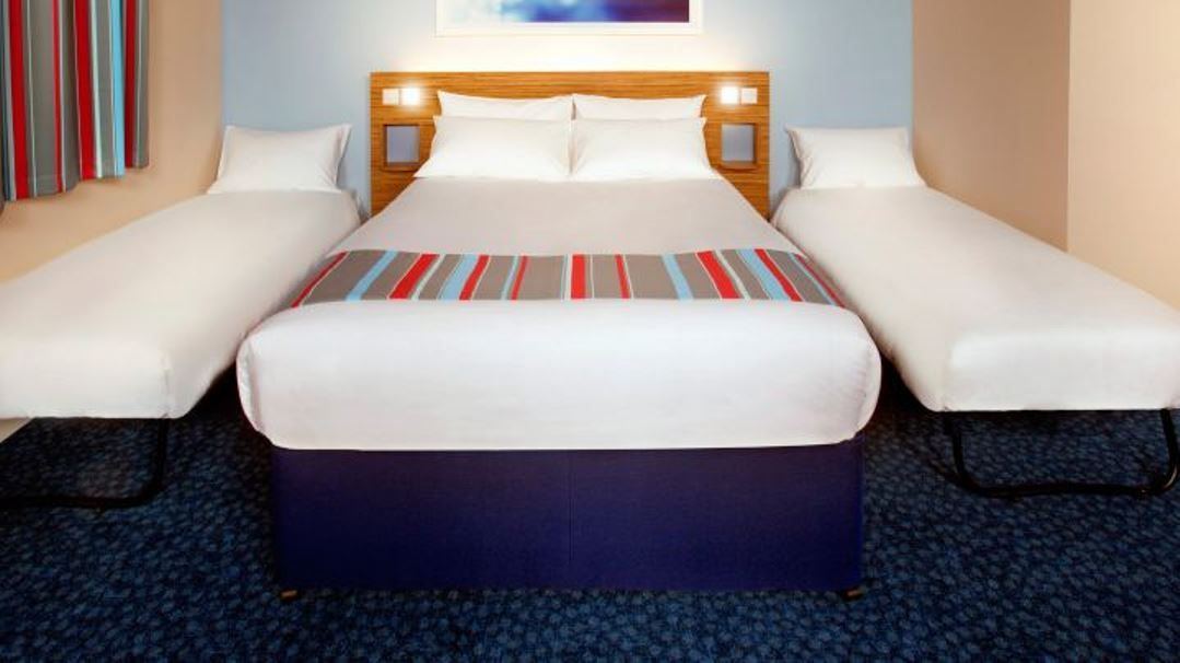 A hotel room with a large bed, two single beds, a wooden headboard, and a blue carpet.