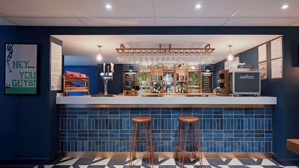 A bar area with two stools, a tiled backsplash, and a variety of bottles and glasses on shelves. - Travelodge Edinburgh Central (Edinburgh)