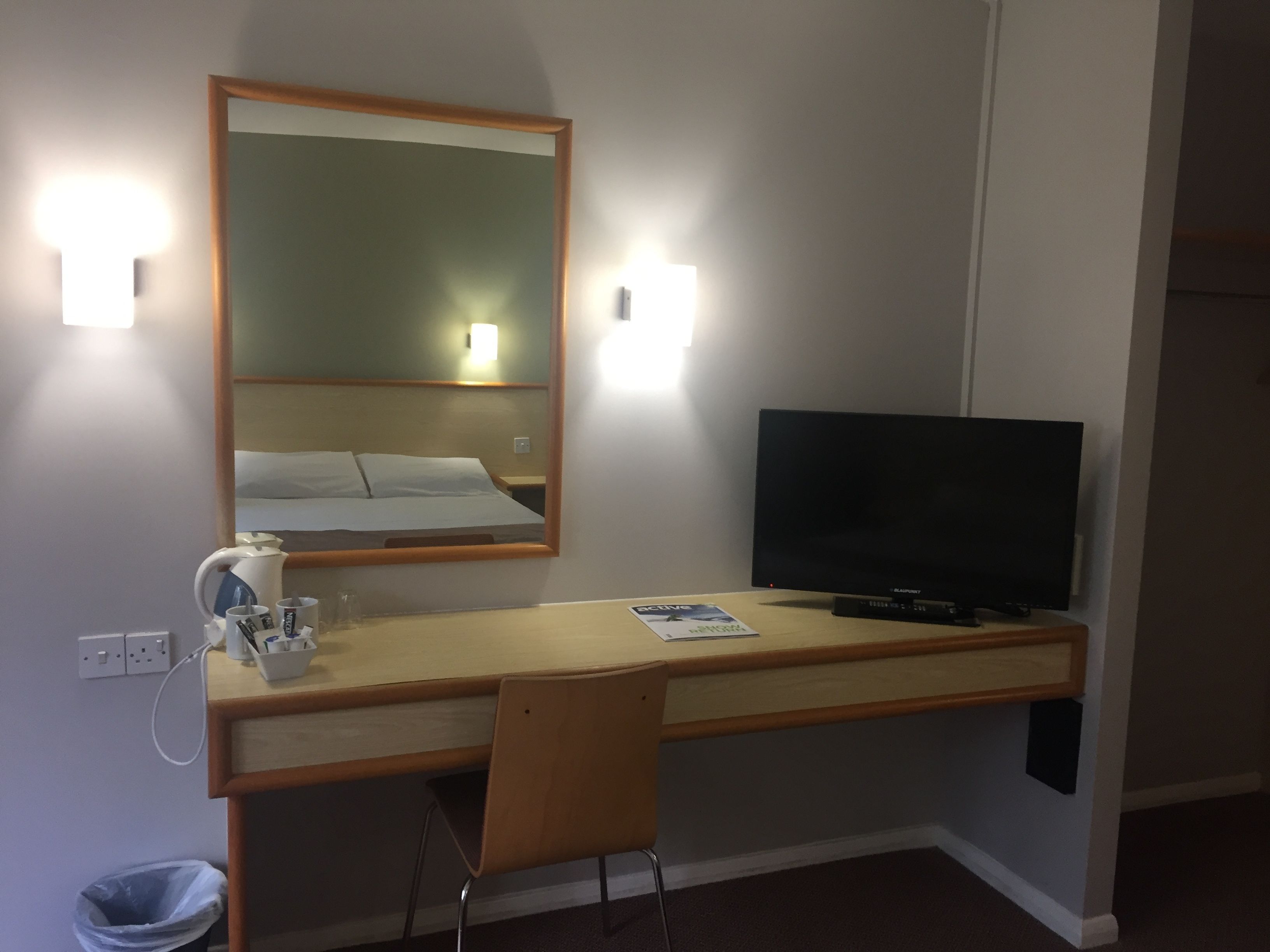 Habitación de hotel con un escritorio de madera, televisor, espejo y cama con ropa blanca.