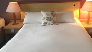 Una cama bien hecha con sábanas blancas y un cojín decorativo, flanqueada por dos lámparas de noche sobre mesitas de madera.
