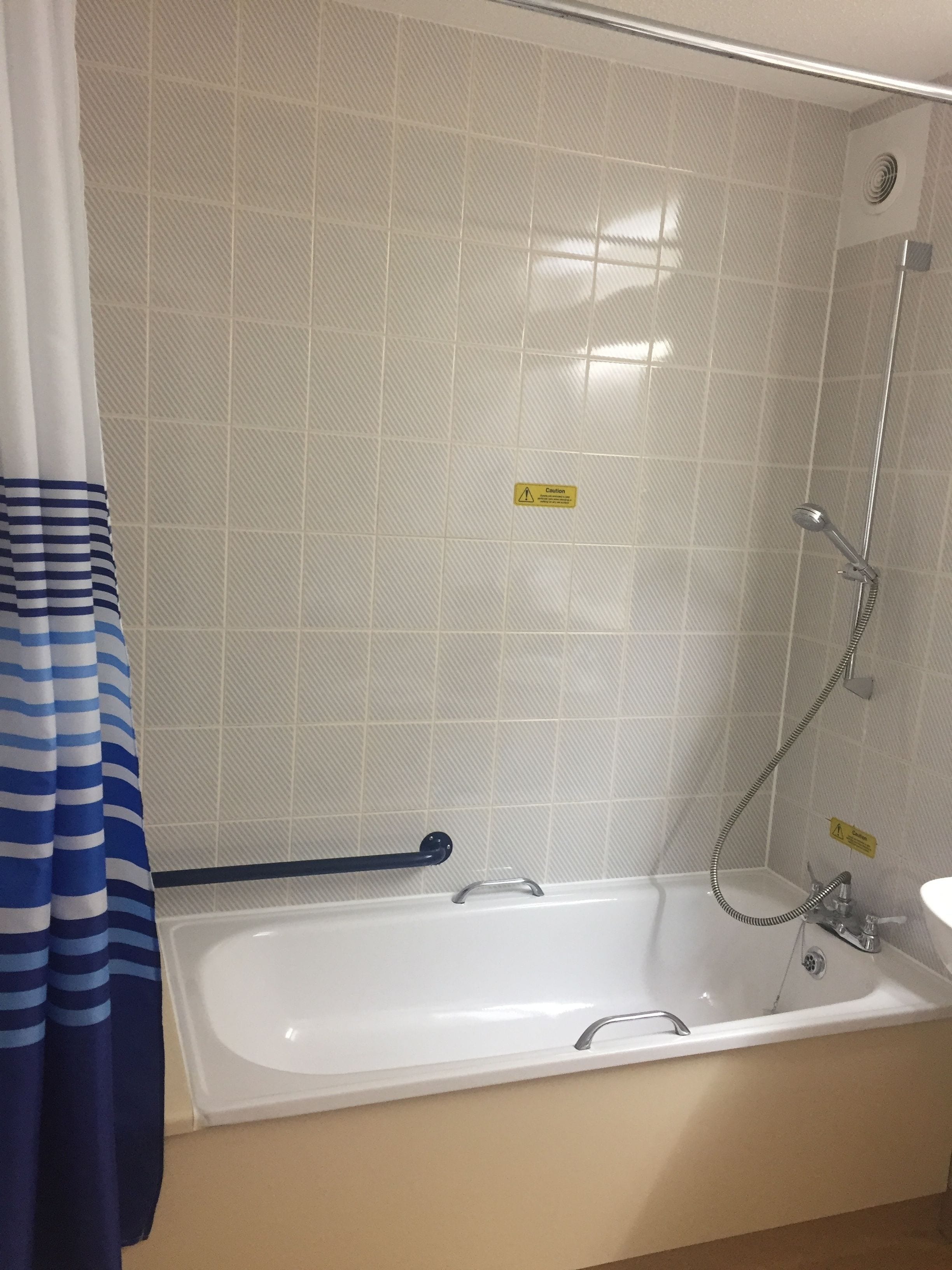 A bathroom with a bathtub, a shower, and a sink. There is a caution sign on the wall and a striped curtain.