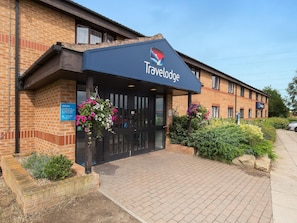 Un hôtel Travelodge avec une façade en briques, un auvent noir à l’entrée et des jardinières suspendues remplies de fleurs.
