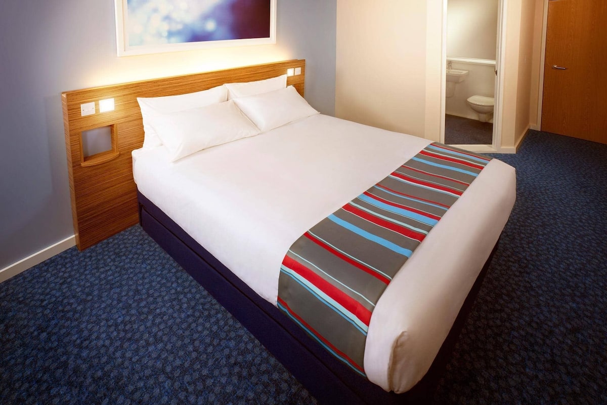 Un lit bien fait, avec du linge de lit blanc et une couverture rayée aux couleurs vives, une tête de lit en bois et un sol recouvert d’une moquette bleue.