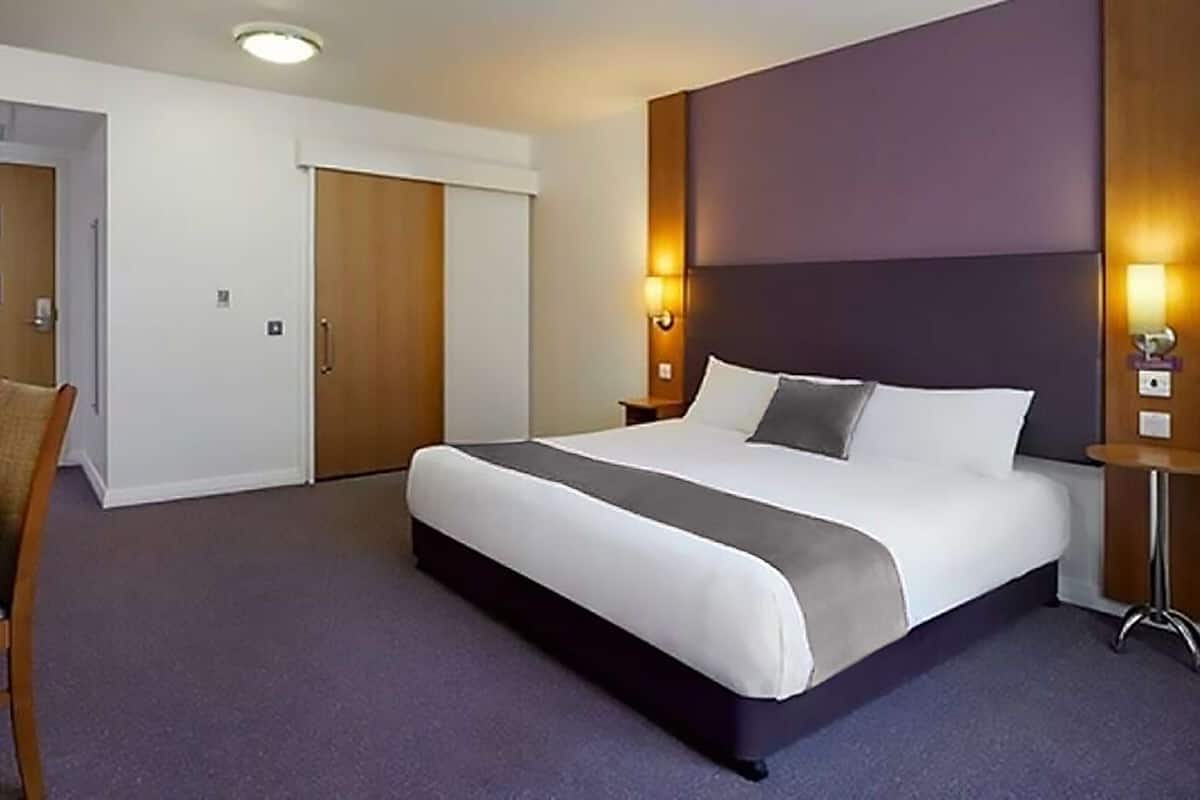 A hotel room with a large bed, a desk, and a chair. There is a door and a wall-mounted light fixture.