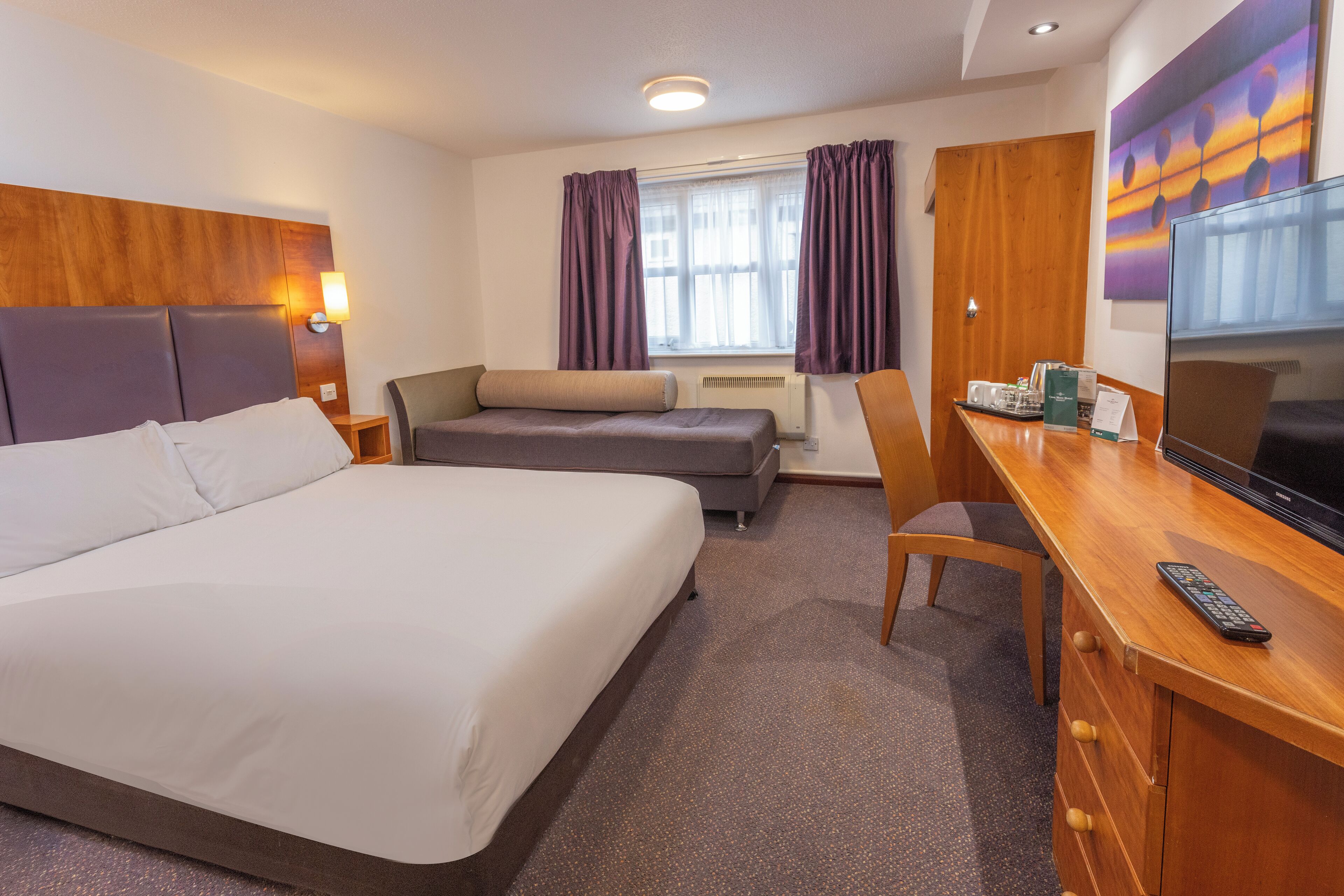 A hotel room with a large bed, a sofa, a desk with a TV, and a window with curtains.