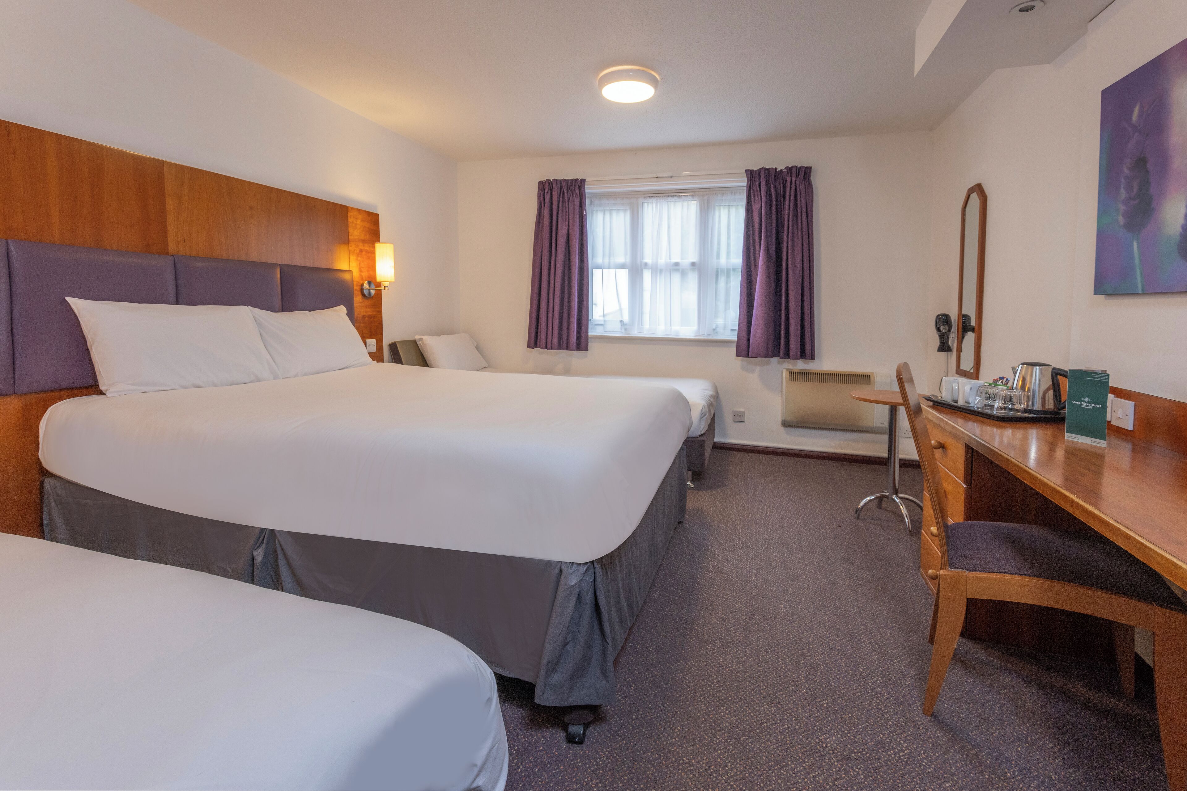 A hotel room with two beds, a desk, a chair, a window with curtains, a radiator, and a painting on the wall.