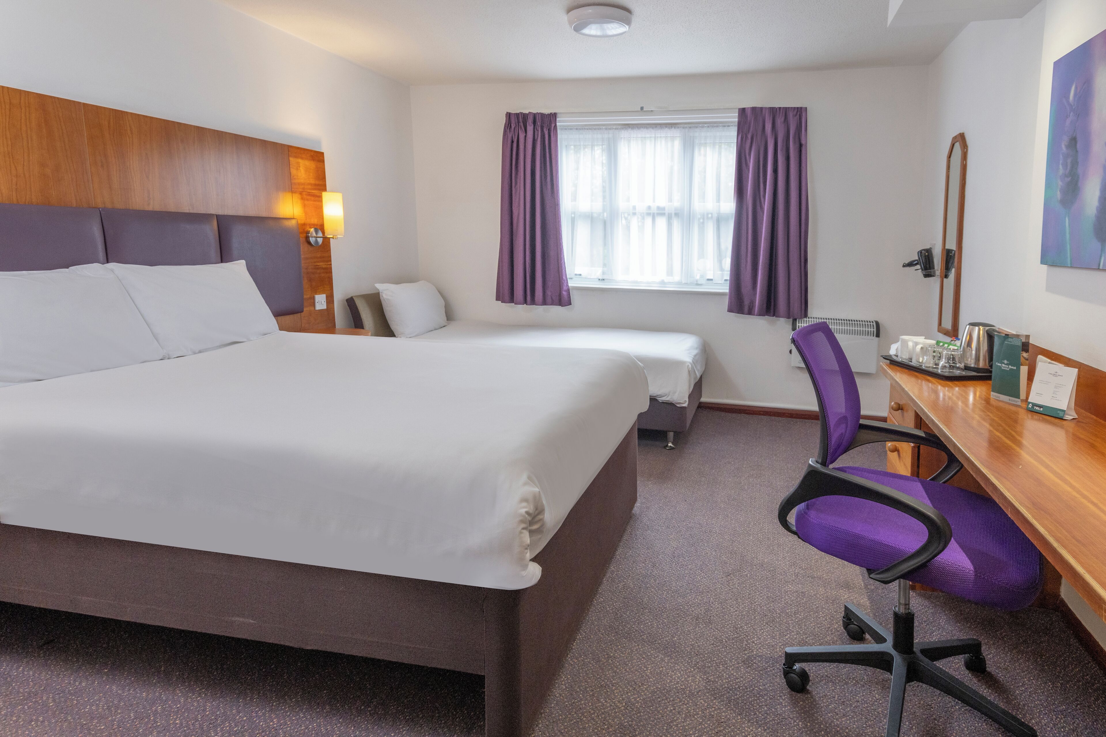 A hotel room with a large bed, a smaller bed, a desk with a chair, a window with purple curtains, and a painting on the wall.