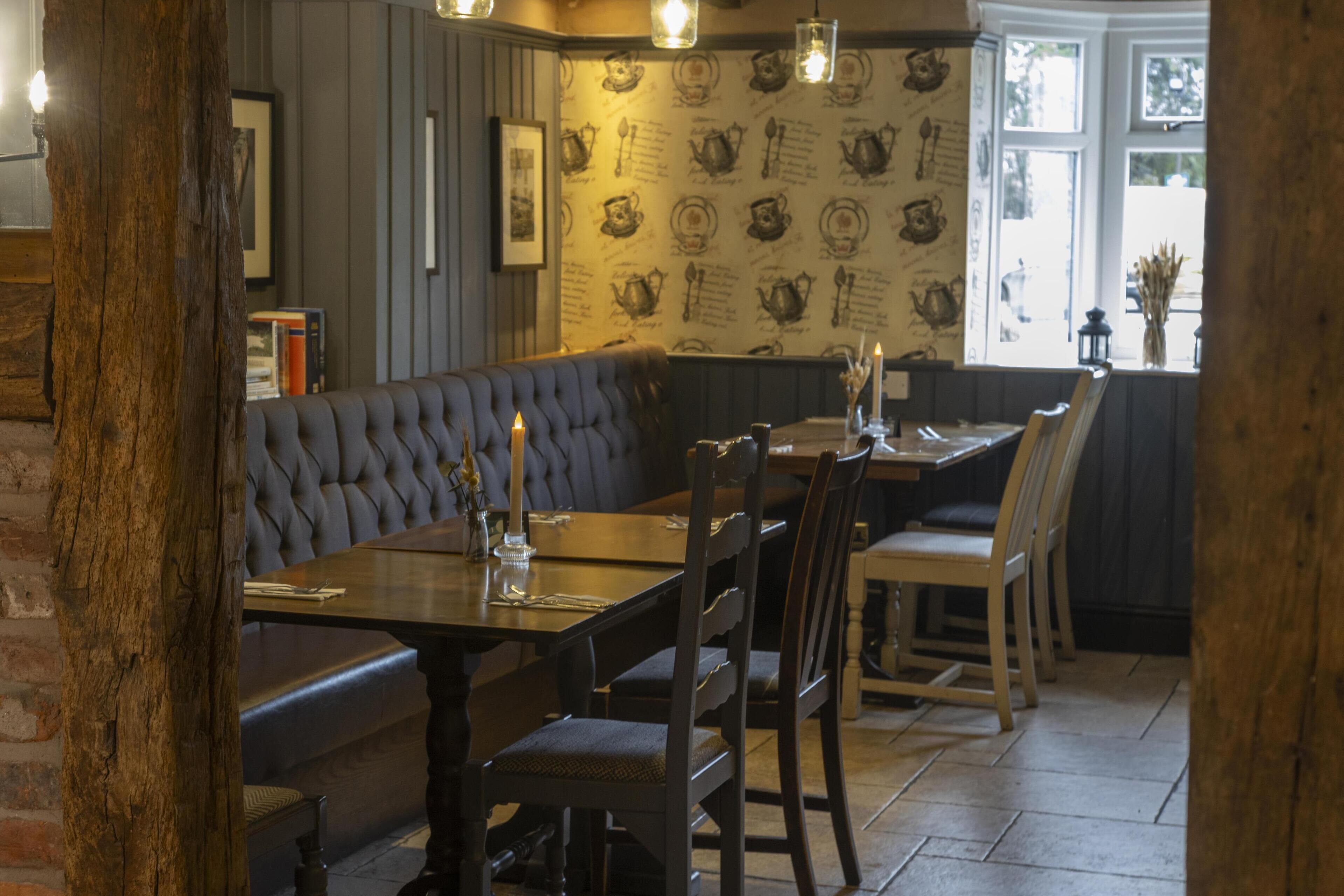 Interior acogedor de un restaurante con una zona de estar con bancos de madera, una mesa alta con sillas y un papel tapiz estampado en la pared de fondo.