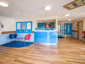 A reception area with a blue counter, a digital screen, a fire extinguisher, and a vending machine. - Travelodge Stafford M6 (Stone)