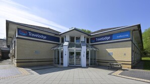 A Travelodge hotel entrance with a clear glass door, a bench outside, and a tiled pavement. - Travelodge Stafford M6 (Stone)