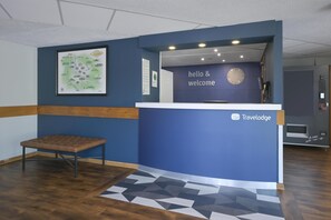 A reception desk with a 'hello & welcome' sign, a framed map on the wall, and a bench in the corner. - Travelodge Stafford M6 (Stone)