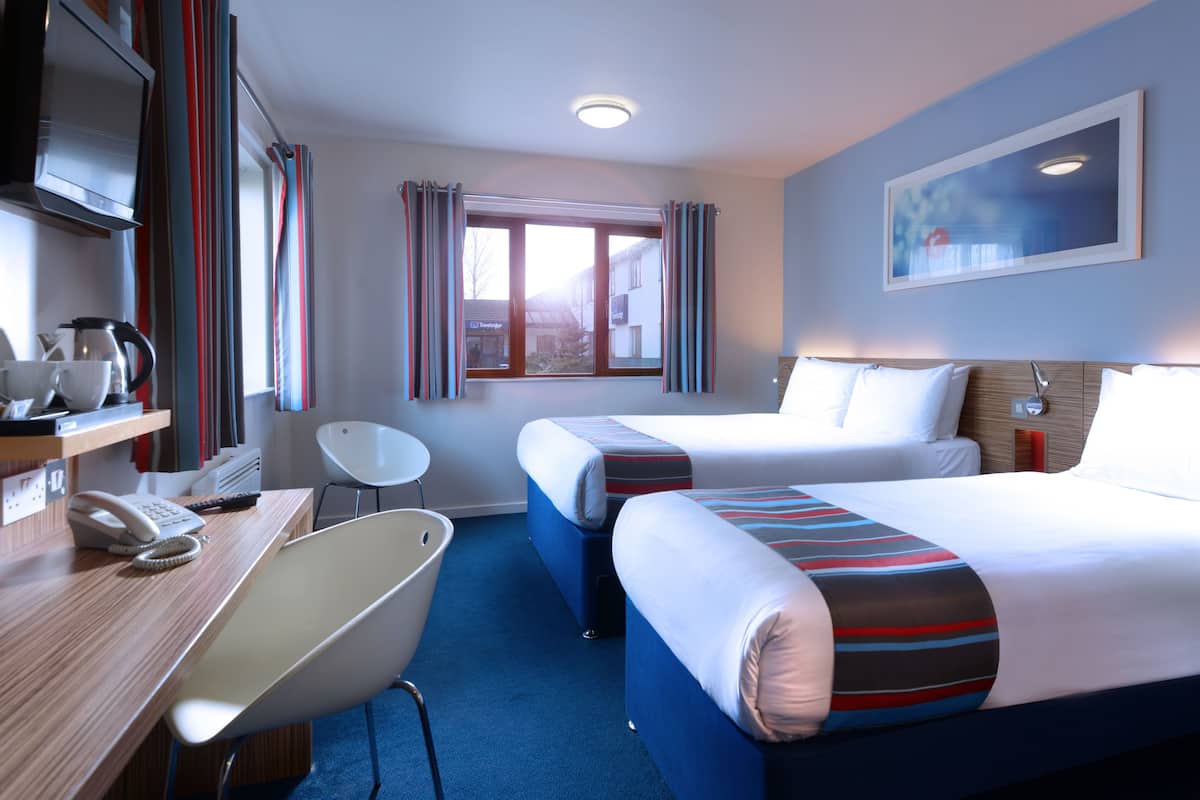 A hotel room with two beds, a desk with a telephone, a chair, and a window with curtains.