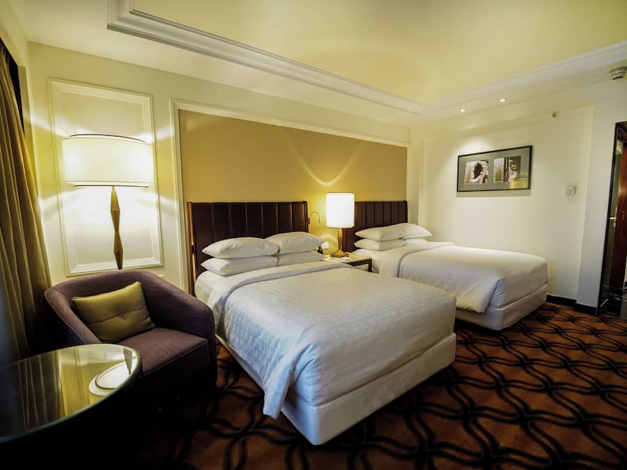 A hotel room with two beds, a chair, a table, and a lamp.