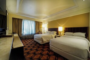 Habitación de hotel con dos camas, un escritorio, un televisor y un ventanal grande con cortinas.