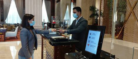 A hotel reception area with a digital screen displaying health and safety guidelines, a man and a woman interacting at the reception desk, and a luxurious interior with marble flooring and elegant decor.