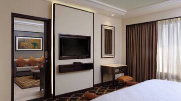 A hotel room with a large bed, a flat-screen TV mounted on the wall, a desk with a chair, and a sofa with cushions.
