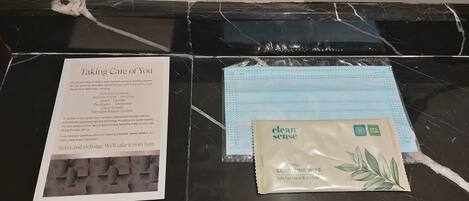 A hotel room with a black marble table, a 'Taking Care of You' pamphlet, a blue face mask, and a 'clean sense' sanitizing wipe packet.