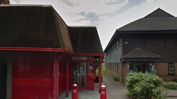 Un bâtiment rouge avec une pancarte « BIENVENUE », un bâtiment brun avec un toit en tuiles et un chemin menant à l’entrée.