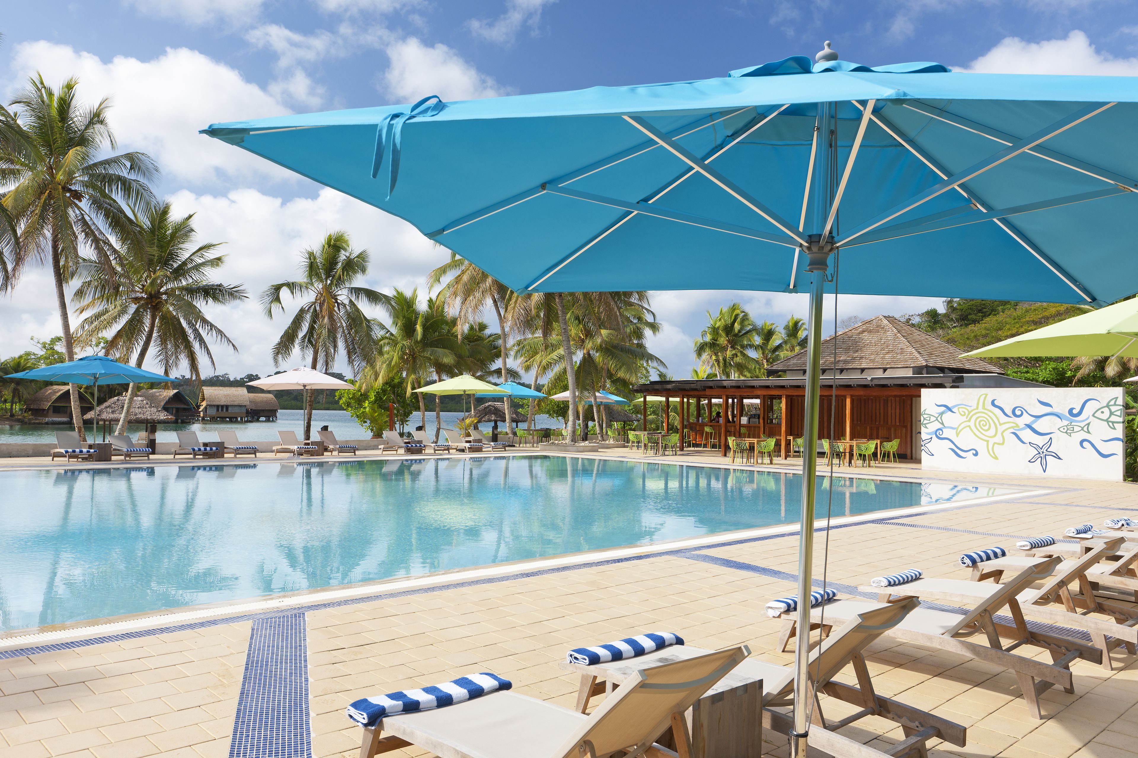 A poolside area with lounge chairs and umbrellas, palm trees, and a thatched-roof structure.
