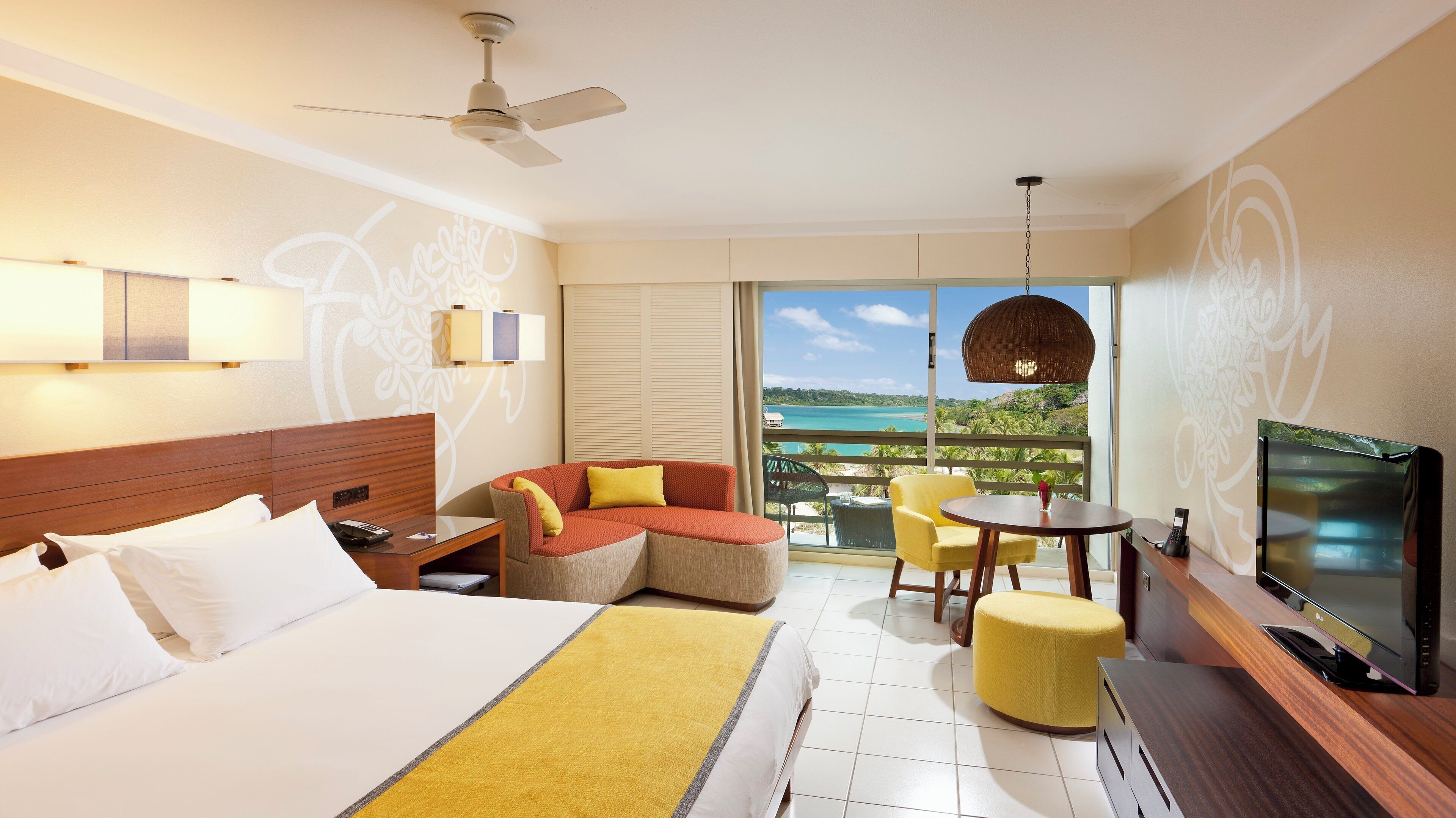 Habitación de hotel con cama, sofá, televisión, balcón con vista al mar y ventilador de techo.