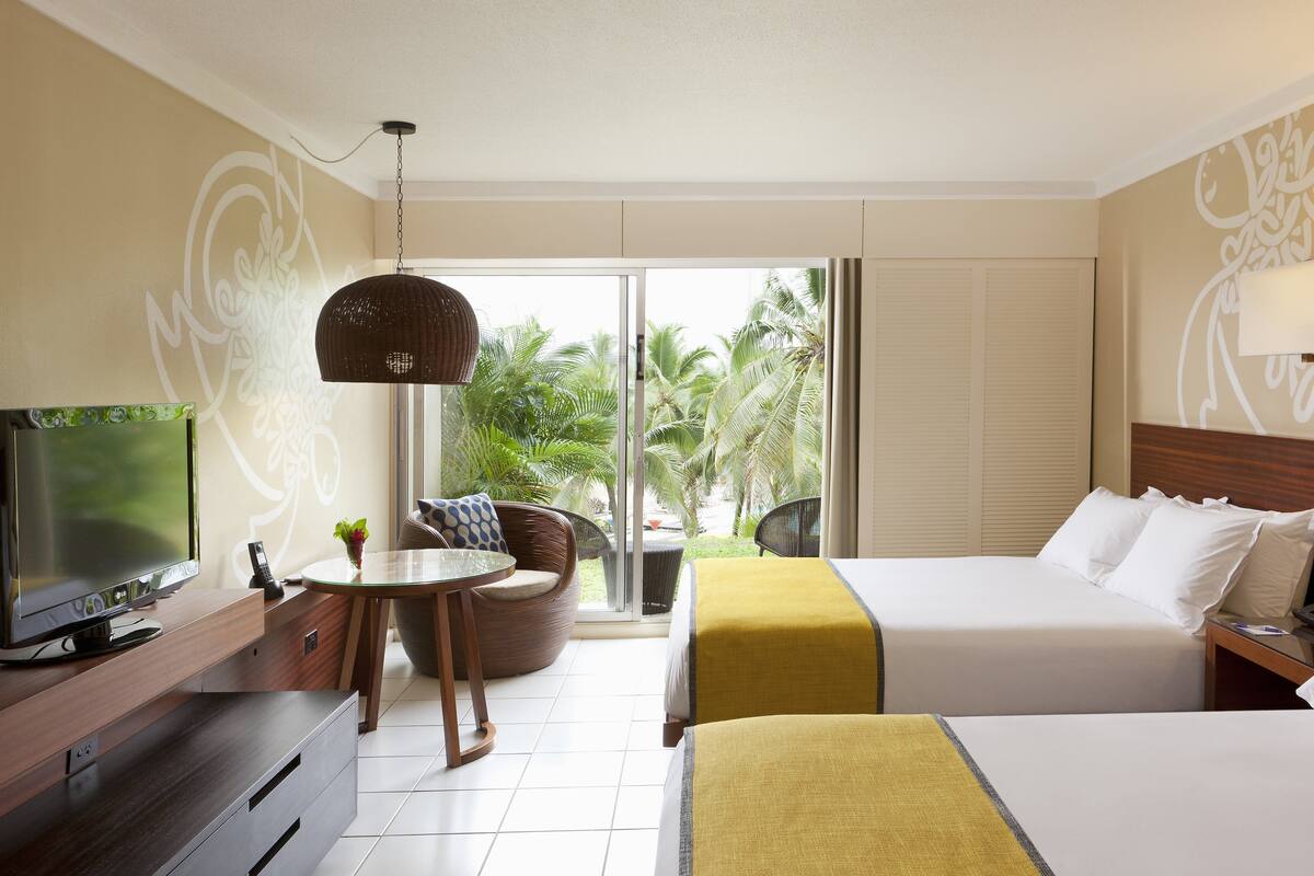 A hotel room with a bed, a TV, a dining area with a table and chairs, and a view of palm trees through the window.
