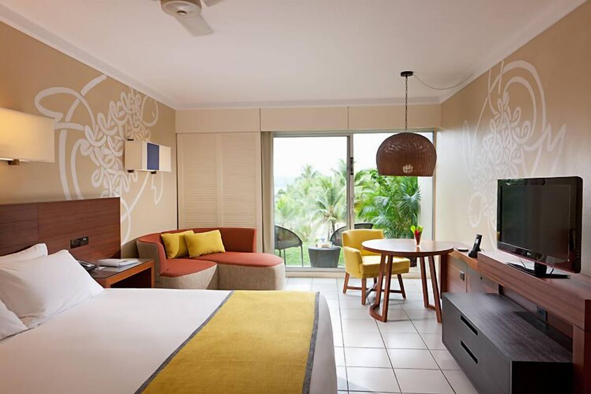 A hotel room with a bed, a sofa, a dining table with chairs, a TV, and a balcony with plants.