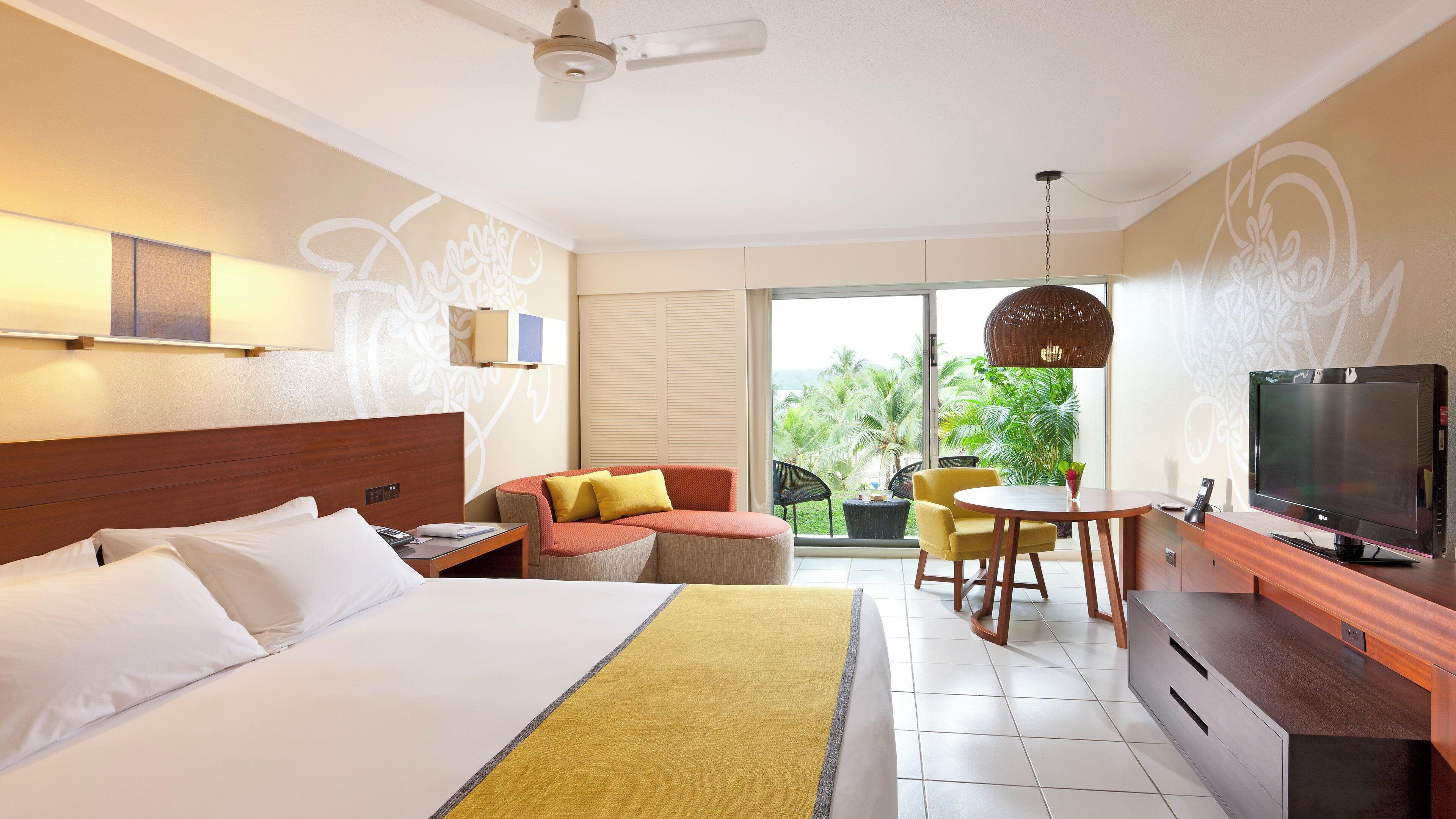 A hotel room with a bed, a sofa, a dining table, a TV, and a balcony with plants.