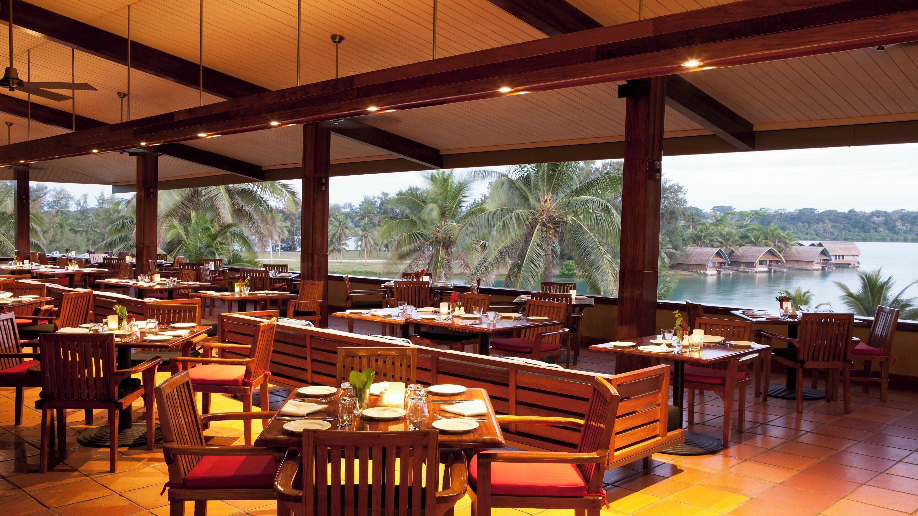 An outdoor restaurant with wooden furniture and tables, overlooking a body of water and palm trees.