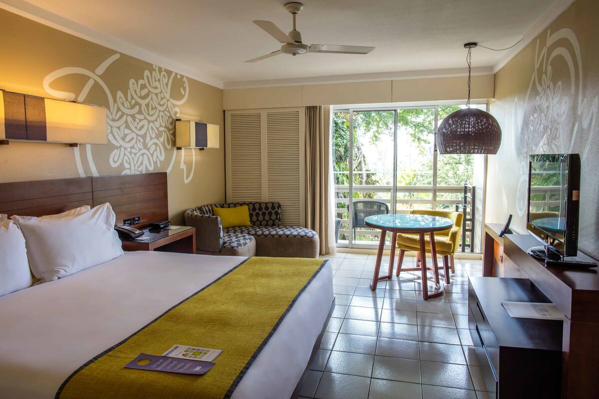 A hotel room with a bed, a desk, a chair, a table, a TV, and a balcony with a view of greenery.