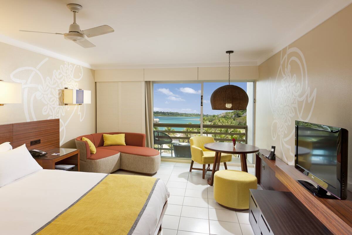 A hotel room with a bed, a sofa, a TV, a balcony with a view of the sea, and a ceiling fan.