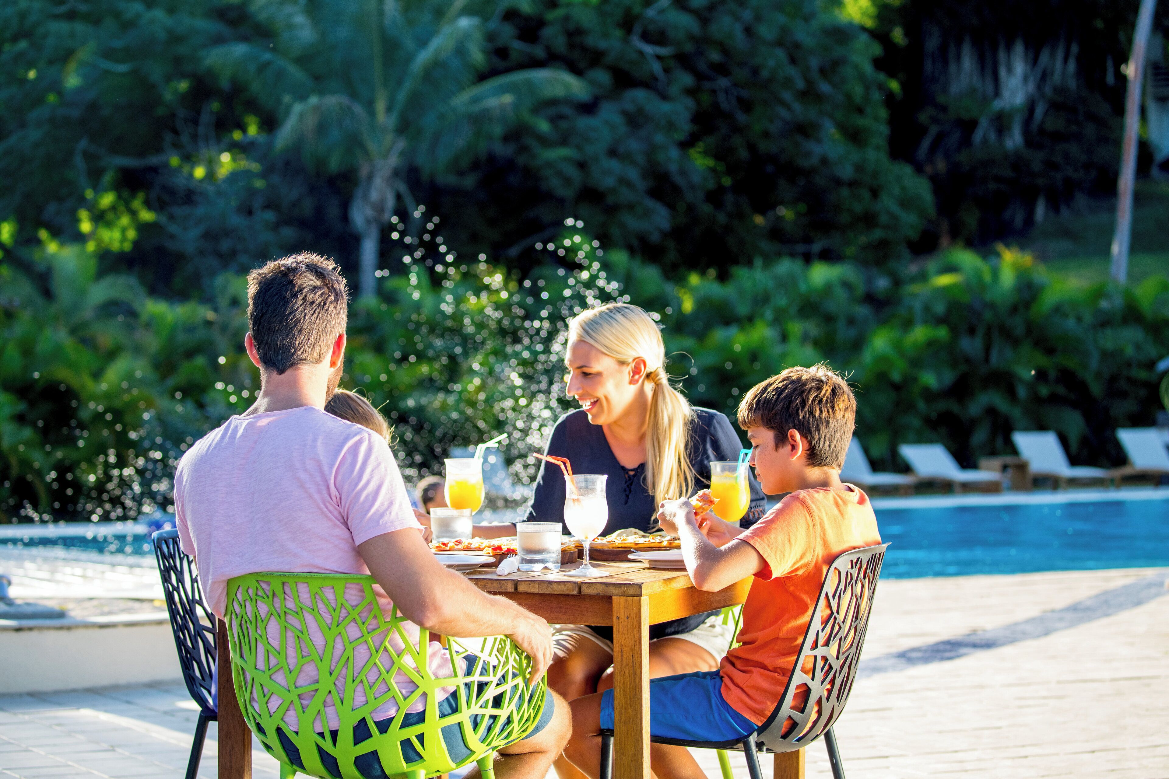 A family dining at an outdoor table by a pool, with chairs and a table set for a meal.