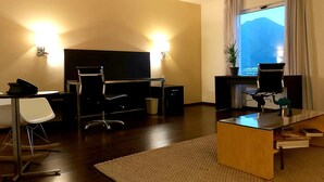 A hotel room with a flat-screen TV, a desk with a computer, a chair, a small table with a plant, and a view of mountains through the window.