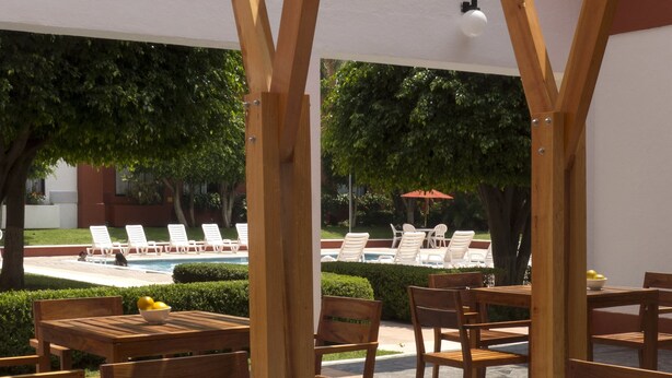 An outdoor wooden pergola with tables and chairs, a pool, and lounge chairs in the background. - Fiesta Inn Leon (León)