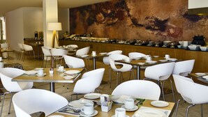 A dining area with white chairs, tables set with plates, cups, and cutlery. A buffet line is visible in the background. - Fiesta Inn Leon (León)