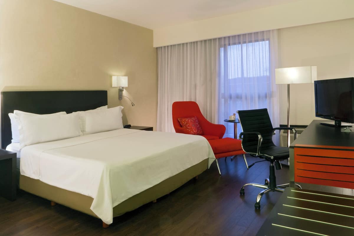 A hotel room with a large bed, a desk with a computer, a red chair, and a television.