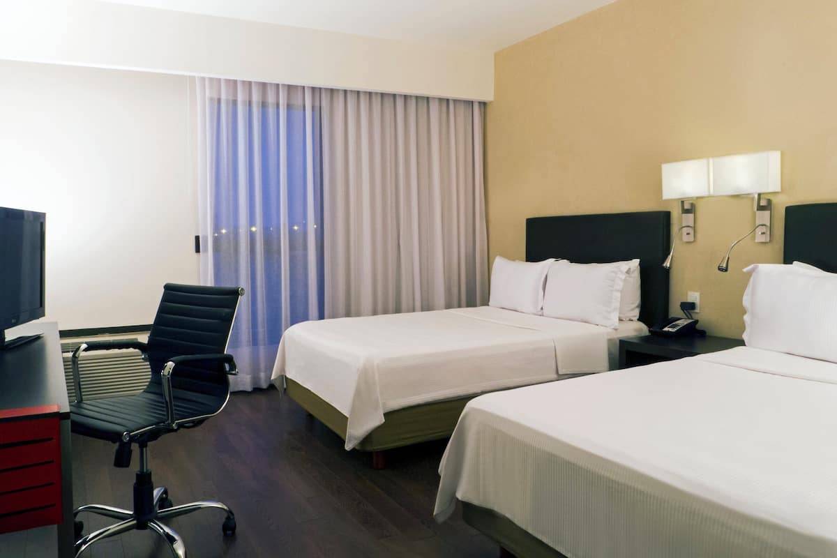 A hotel room with two beds, a desk, a chair, and a large window with curtains.