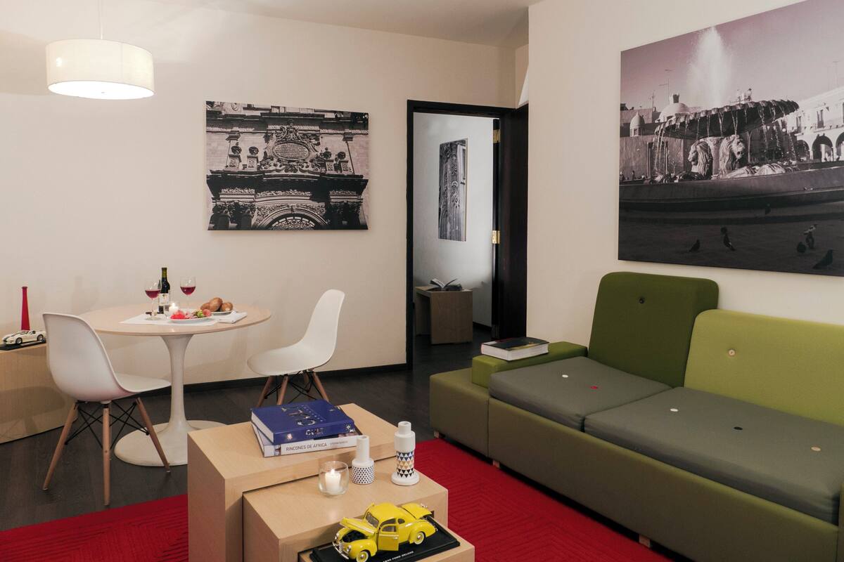A modern living room with a green sofa, a white dining table with chairs, a small coffee table with books and a toy car, and two wall-mounted paintings.