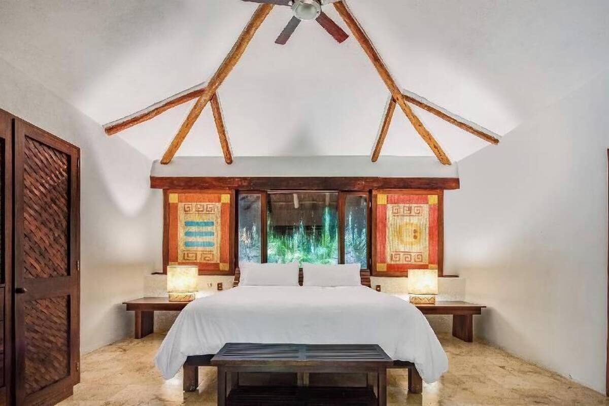 A bedroom with a bed, two bedside tables, a ceiling fan, and a view of greenery through the window.